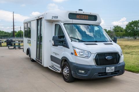 DCTA's white ADA-accessible "Access" bus sits on the pavement, with blue sky, grassy grounds, and picnic tables behind it.